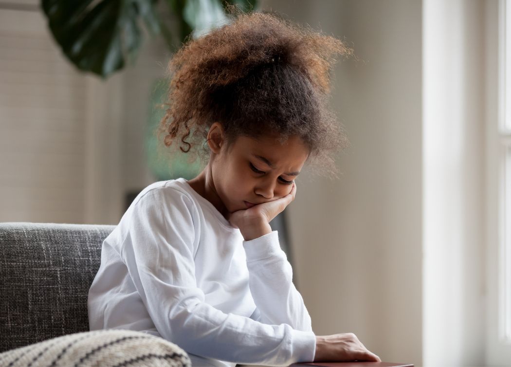 Young girl sitting with head in hand, looking down thoughtfully or sadly indoors.