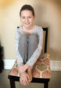 Sara Smiling girl sitting on chair with knees hugged to chest, posing against a plain wall.