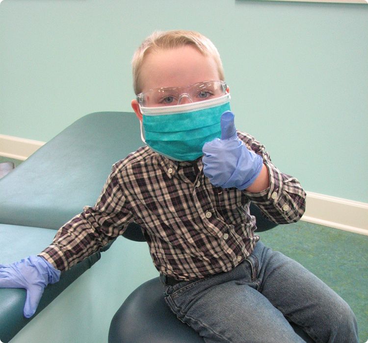Young boy wearing mask, goggles, and gloves gives thumbs up while sitting on exam table.