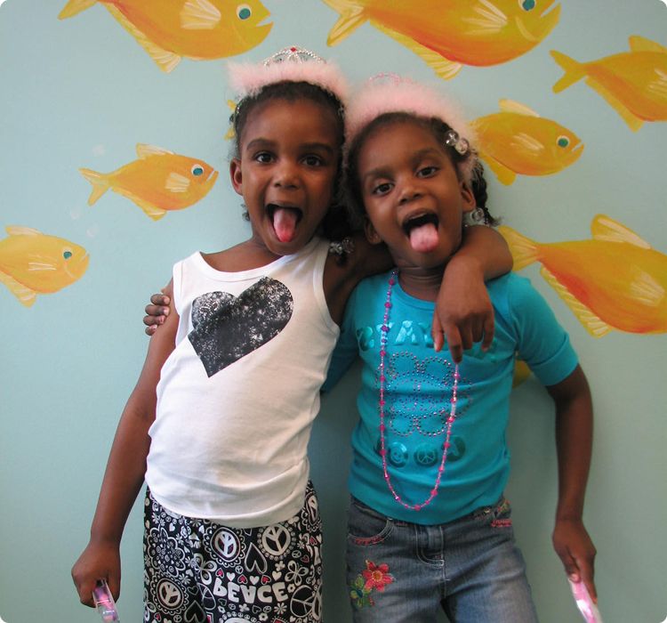 Sedation dentistry page content image 2 Two girls wearing tiaras hug and stick out their tongues in front of a fish-decorated wall.