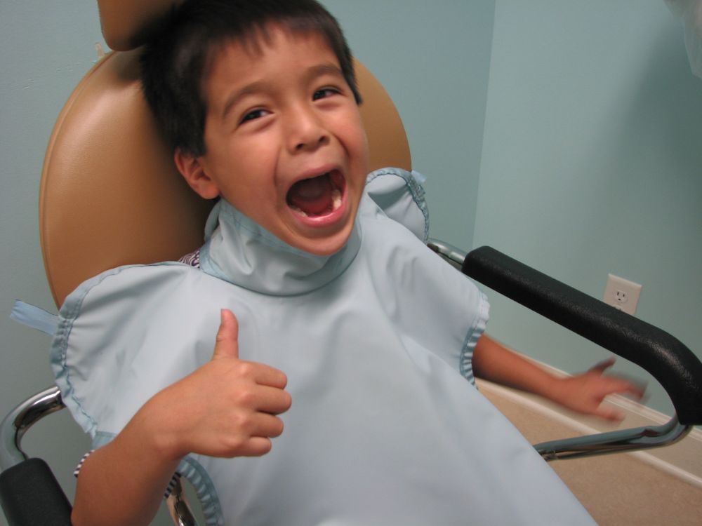 Boy in dental chair giving thumbs up with mouth open, showing bravery during exam.