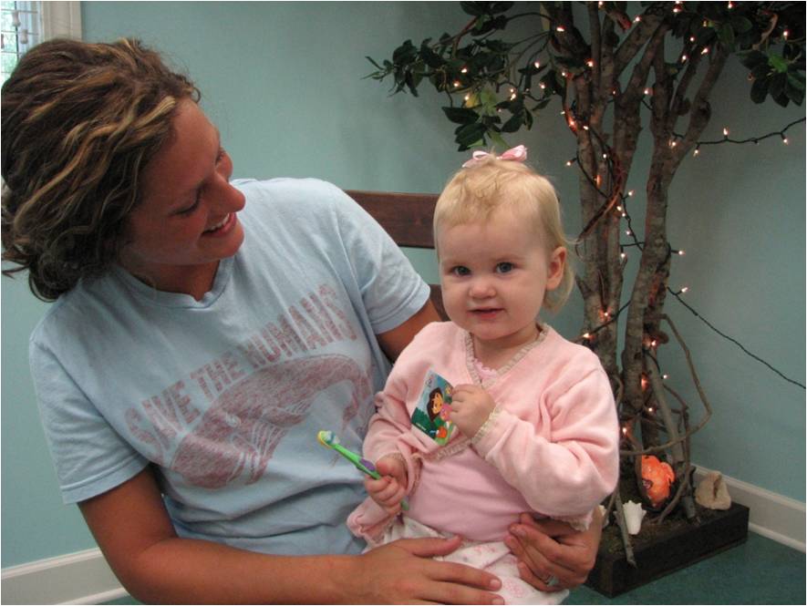 Testimonials page content image 2 Mother holding toddler with toothbrush in a dental office, showing positive early dental visit.