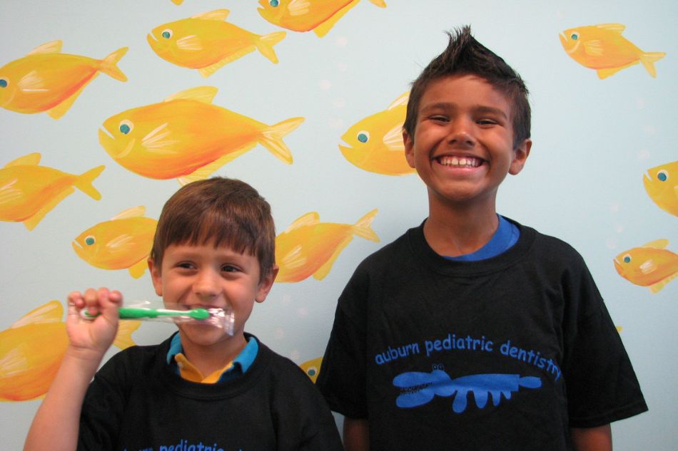 Two boys, one brushing teeth, both looking at the camera and smiling