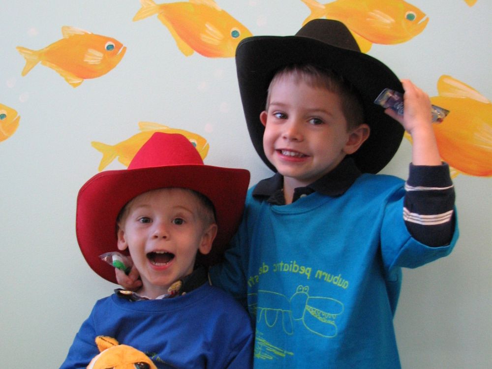 Two young boys in cowboy hats smiling in front of a wall with bright fish decorations.