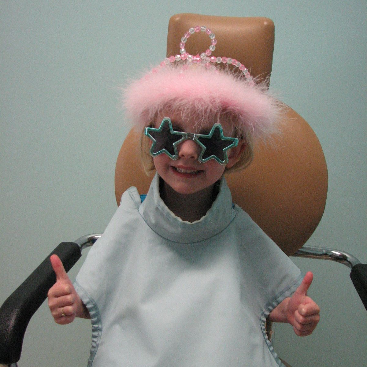 Child in dental chair wearing star-shaped glasses and pink tiara gives thumbs up, showing comfort at visit.