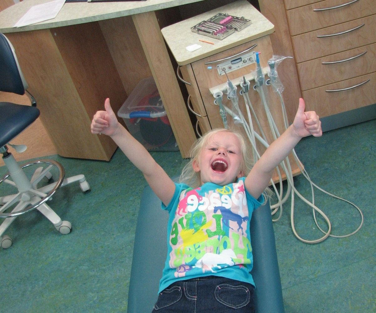 Child in dental chair with arms raised and thumbs up, showing excitement and positive dental visit.